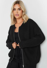 Svart ribbad cardigan med långa ärmar och dragkedja fram. Tillverkad av strukturerat stickat tyg, med avslappnad passform och öppen halsringning.