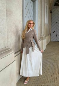 Femme aux longs cheveux blonds portant une veste beige boutonnée et une longue jupe blanche, debout près d’un mur texturé et de portes décoratives.