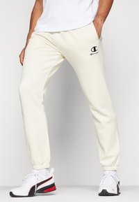 Pantalons de jogging crème avec une taille élastique, des ourlets resserrés et un petit logo Champion noir sur la cuisse gauche. Texture de tissu douce.