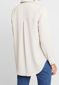 Blouse à manches longues de couleur crème, avec col, détail pli au dos et poignet boutonné, fabriquée dans un tissu doux et texturé. Ourlet arrondi.