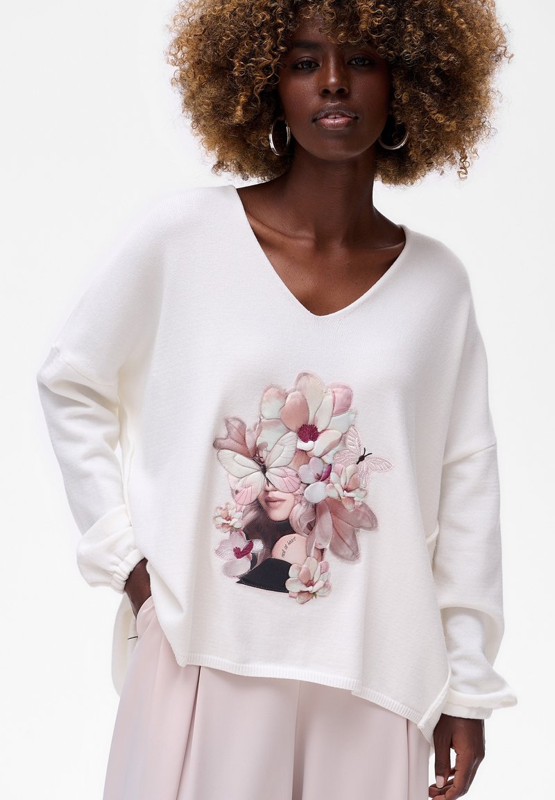 Vrouw met krullend haar draagt een witte V-halstrui met bloem- en vlinderkunst, gecombineerd met lichtroze wijde broek.