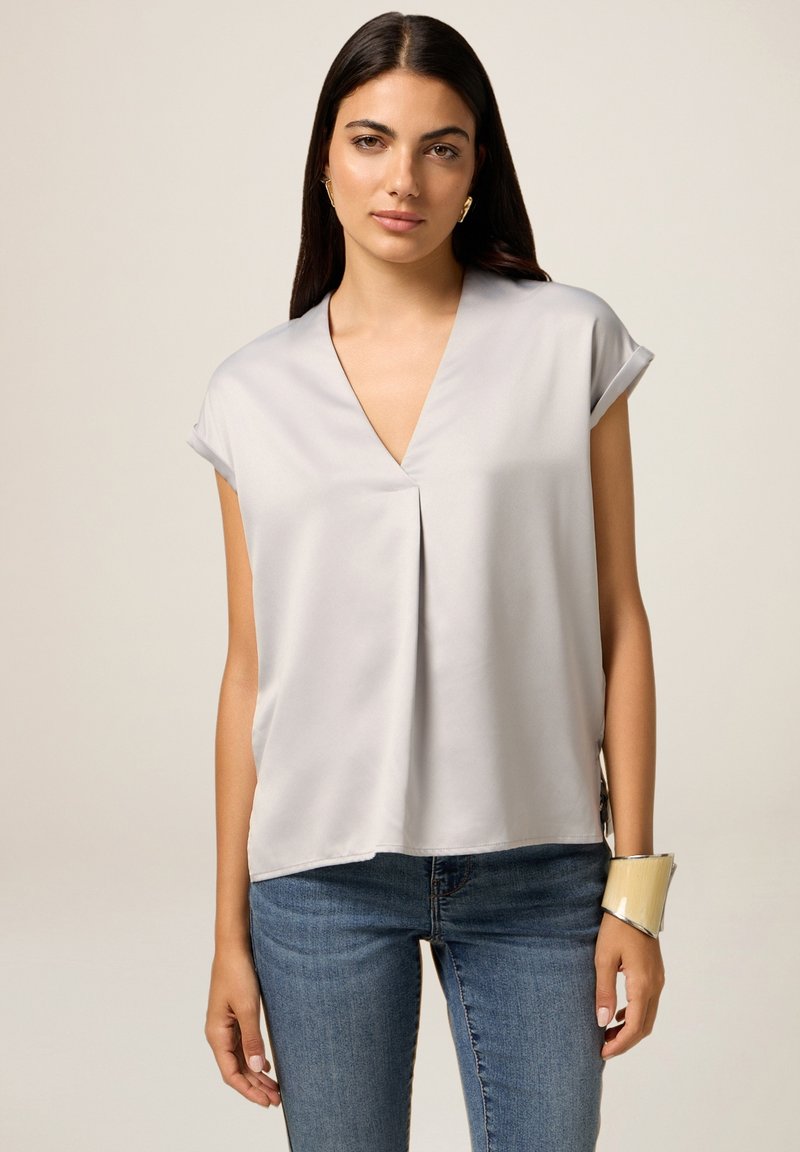 Lys grå kortærmet bluse med V-hals og plisseret front. Glat tekstur og løs pasform, sat sammen med blå denim jeans og et armbånd.