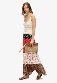 Gehaakte tanktop in crème, gelaagde maxi rok met paisleypatronen in rood en zwart, bruine geweven clutchtas, bronzen sandalen met cirkelvormige accenten.