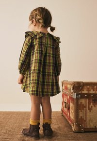 Karo-Kleid in Braun, Grün und Lila mit Rüschensaum und Knopfverschluss. Braune Stiefel mit gelben Socken ergänzen den Look.