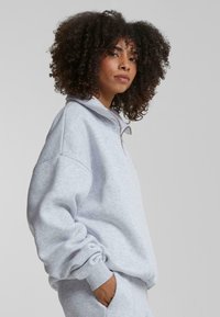 Lys grå hettejakke med glidelås og høy krage, ribbestrikkede mansjetter og avslappet passform. Myk fleece-materiale med en glatt tekstur.