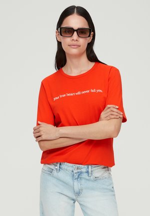 T-shirt en coton rouge à manches courtes, avec un texte blanc "ton vrai cœur ne te laissera jamais tomber" sur le devant. Porté avec un jean bleu clair.