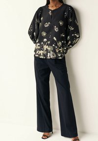 Schwarze Bluse mit floralem Muster in Grün und Weiß, mit einem runden Ausschnitt und elastischen Puffärmeln, kombiniert mit marineblauen weiten Hosen.