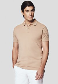 Beżowa koszulka polo z krótkim rękawem i klasycznym kołnierzykiem, z zapięciem na dwa guziki i miękką fakturą. Noszona z białymi szortami dla kontrastu.