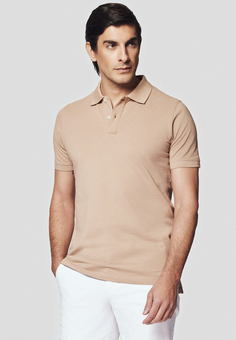 Beżowa koszulka polo z krótkim rękawem i klasycznym kołnierzykiem, z zapięciem na dwa guziki i miękką fakturą. Noszona z białymi szortami dla kontrastu.