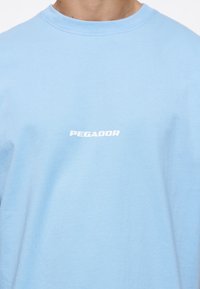 Lyseblå bomulls t-skjorte med crew neck og korte ermer. "PEGADOR" er trykt i hvitt over brystet. Enkelt design, myk tekstur.