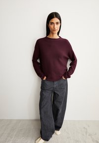 Bordeauxfarbener Pullover mit Rundhalsausschnitt, kombiniert mit grauen, weitgeschnittenen Jeans und hellen Schuhen. Beide Kleidungsstücke haben einen lockeren Schnitt.