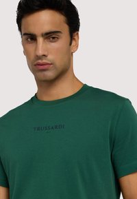 T-shirt verde in cotone con maniche corte, caratterizzata da un collo rotondo e una stampa del logo blu scuro con la scritta "TRUSSARDI 1911" sul petto.