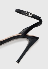 Sorte højhælede sandaler med tynd ankelrem, der har en sølv spænde, og en slank stiletto hæl på en ensfarvet baggrund.