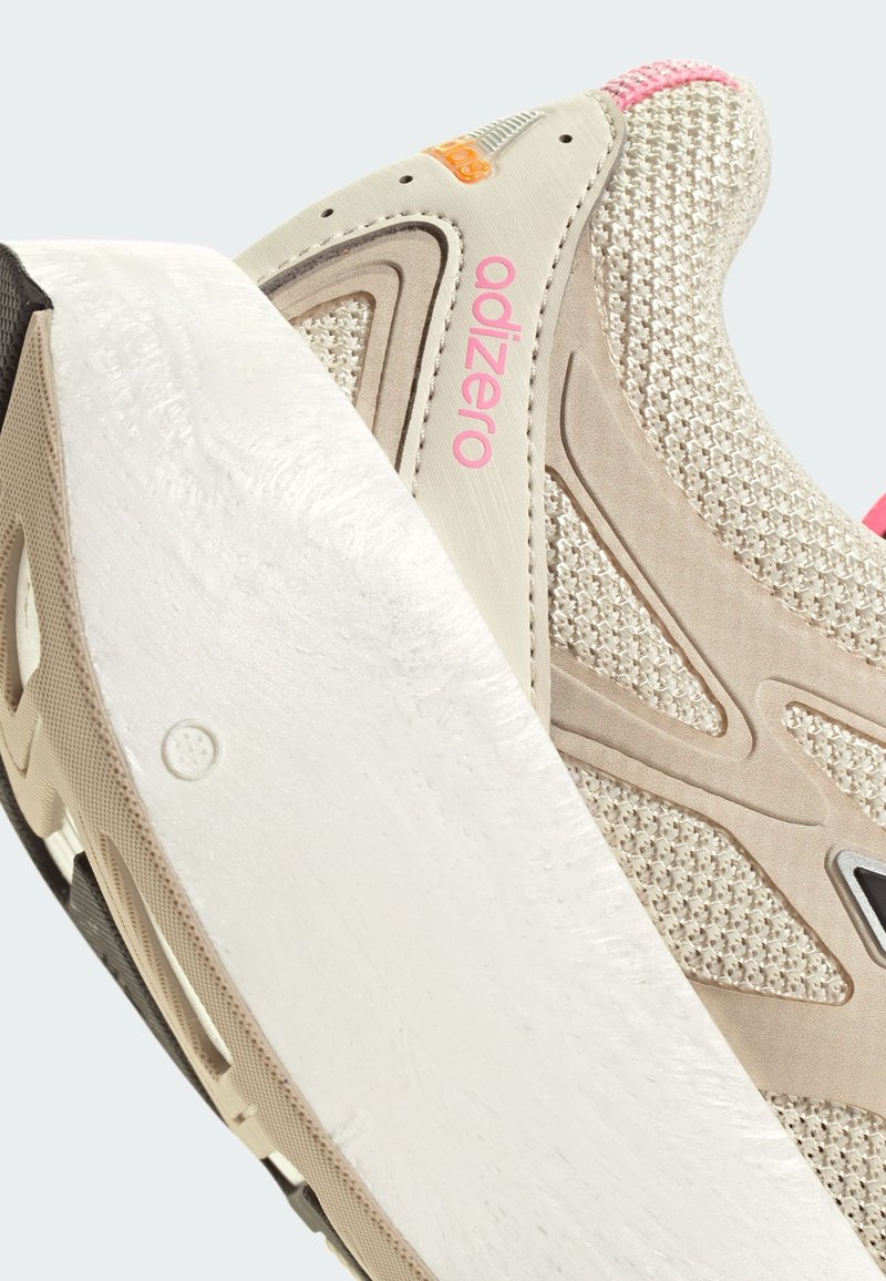 Beigefarbener Sportschuh mit einer strukturierten Oberseite, weißer Sohle und pinken Akzenten. Verfügt über ein Logo und ein gestuftes Design an den Seiten.