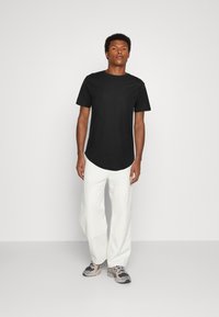 Schwarzes kurzärmliges T-Shirt mit abgerundetem Saum, kombiniert mit locker sitzenden weißen Hosen und grauen Sneakers. Schlichtes Design, weicher Stoff.