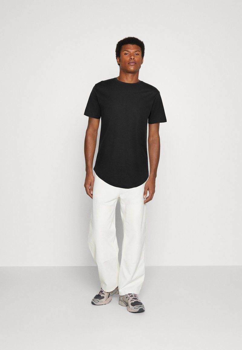 Schwarzes kurzärmliges T-Shirt mit abgerundetem Saum, kombiniert mit locker sitzenden weißen Hosen und grauen Sneakers. Schlichtes Design, weicher Stoff.