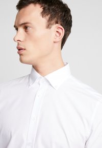 Weißes Hemd mit Button-Down-Kragen, glatter Textur und sichtbaren Knöpfen entlang der Vorderseite. Minimales Design, klare Linien.