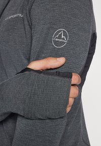 Dark gray long-sleeve garment with textured fabric, featuring a contrasting black cuff and a white logo on the upper arm.