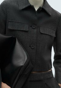 Czarna, kraciasta marynarka z dwoma kieszeniami na piersi i zapięciem na guziki, zestawiona z gładką, czarną skórzaną torebką. Krótki, dopasowany fason.