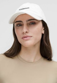 Bijela bejzbol kapa s "lululemon" logotipom u crnoj izvezenoj boji, nosi se uz glatku, svijetlu bež majicu s okruglim izrezom.