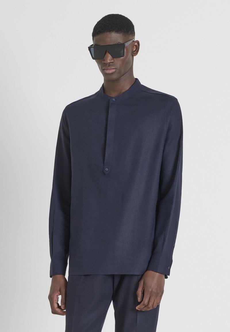 Camicia a maniche lunghe blu navy con colletto alla cinese, pattina con un solo bottone e tessuto strutturato. Indossata con occhiali da sole neri angolari.