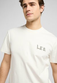 Valkoinen puuvillat-paita, jossa on lyhyet hihat ja harmaa "LEE" -logo painettuna vasempaan rintapieleen. Silkkinen rakenne.