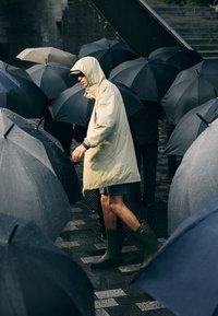 Bežový pršiplášť s kapucňou, tmavé šortky a zelené gumové čižmy, obklopený čiernymi dáždnikmi na mokrej dlažobnej ceste.