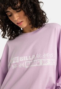 Purple cotton long-sleeve shirt featuring a white graphic logo that includes floral and sun motifs. Soft texture, relaxed fit, crew neckline.