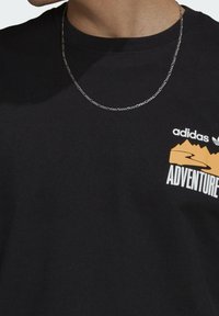 Czarna bawełniana koszulka z krótkim rękawem, z białym i pomarańczowym graficznym napisem "adidas ADVENTURE" oraz srebrnym naszyjnikiem łańcuszkowym.