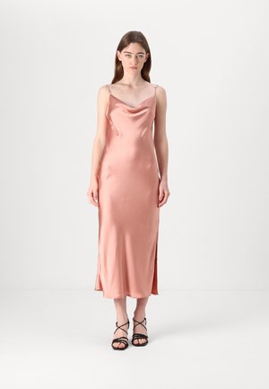 Satin slip dress in soft pink with a cowl neckline, thin straps, side slits, and a smooth texture. Paired with black strappy sandals.