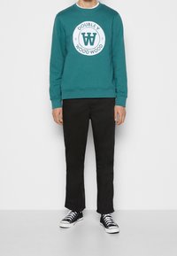 Tyrkysová mikina s kulatým bílým logem a textem, doplněná černými kalhotami a černobílými teniskami. Jednoduché pozadí.