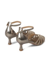 Tacones metálicos plateados con un tacón bajo y acampanado. Presentan un diseño con correas adornadas con pequeñas gemas transparentes. Textura suave y estilo de punta abierta.