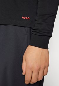 Blízký záběr ruky osoby vedle manžety černé košile s dlouhým rukávem, na které je červené logo "HUGO", a kapsy tmavě šedých kalhot.