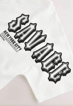 Biele tričko s výrazným čiernym nápisom „SAVAGE NEW YORK CITY“. Dizajn obsahuje komplikovanú, dramatickú typografiu a klasický strih.