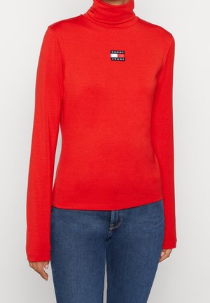 Kobieta ubrana w jasnoczerwoną bluzkę z długim rękawem i golfem oraz małym prostokątnym logo „Tommy Jeans” na piersi, zestawioną z niebieskimi jeansami.