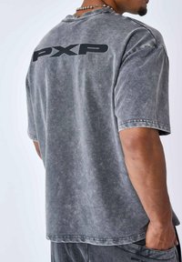 Camiseta de gran tamaño en gris con una textura desvanecida, que presenta una impresión en negro de "PXP" en la parte posterior. Mangas cortas y escote redondo.