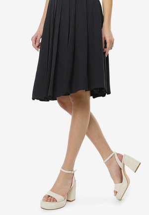 Zapatillas de plataforma blancas con un grueso tacón cuadrado y tira en el tobillo. Material suave y diseño de punta abierta, complementando un vestido negro plisado.