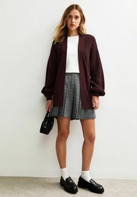 Burgundy knitted cardigan, white fitted top, gray plaid skirt, black loafers, white socks, holding a small black bag, minimalist background.