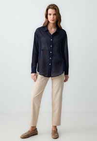 Navy blue, lightweight, button-up shirt with long sleeves and a rounded hem. Paired with beige, straight-leg trousers and brown slip-on shoes.