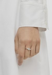 Anillo de plata con una piedra verde claro, con acentos florales. La mano descansa sobre un fondo de tela blanca.