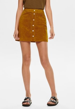 Mujer con minifalda de ante de color mostaza con botones delanteros y bolsillos, top marrón y sandalias negras de doble tira, posando frente a fondo blanco.