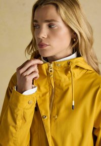 Geltona vandeniui atspari striukė su užtrauktuku priekyje, reguliuojama kepurė ir sagčių rankogaliai. Lygus paviršius su subtiliu ženklinimu.