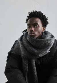 Fiatal férfi göndör hajjal, nagy sz�ürke sálat és fekete steppelt dzsekit viselve, egyszínű világos háttér előtt.