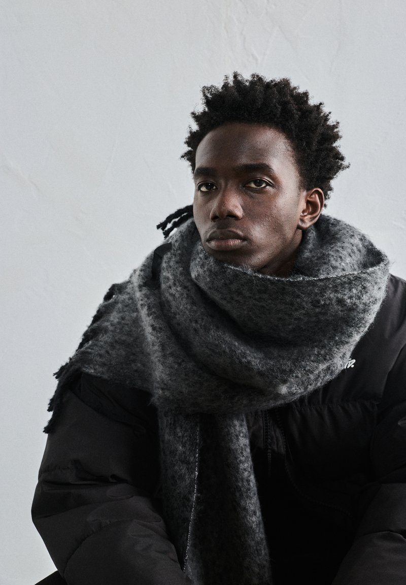 Fiatal férfi göndör hajjal, nagy szürke sálat és fekete steppelt dzsekit viselve, egyszínű világos háttér előtt.