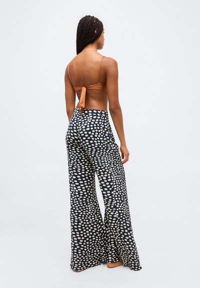 Moteris su pintais plaukais, dėvinti rudą palaidinę, su rišimu nugaros pusėje, ir tamsiai mėlynas platėjančias kelnes su dideliais baltais taškais, stovinti basomis.