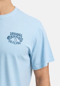 T-shirt en coton bleu clair à manches courtes, avec un graphisme central en texte bleu foncé portant la mention "VOTRE GUÉRISSON ÉNERGÉTIQUE."