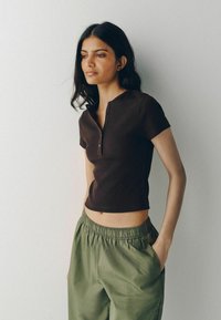 Tânără cu păr lung și închis la culoare, purtând o cămașă neagră cu mâneci scurte, nasturi și pantaloni largi verde-olive, stând cu o mână în buzunar.