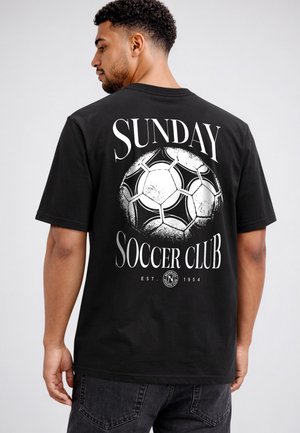 Mann trägt ein schwarzes T-Shirt mit dem Schriftzug "Sunday Soccer Club" und einer Fußball-Grafik auf dem Rücken, dem Kamera abgewandt.