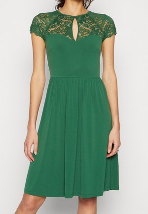 Robe verte mi-longue jusqu'aux genoux avec des manches courtes en dentelle, un décolleté trou de serrure, un corsage ajusté et une jupe froncée portée par une personne debout.