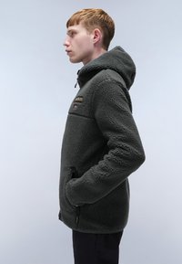 Felpa con cappuccio in pile grigio scuro, con zip frontale, cappuccio regolabile e tasche laterali. Presenta una superficie testurizzata e dettagli del logo.